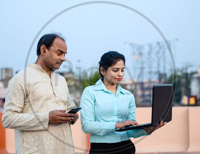Young Indian Female In Formal Dress With Laptop Showing Something To A Middle Aged Man On Internet, The Man Using His Smartphone And Looking On Laptop.