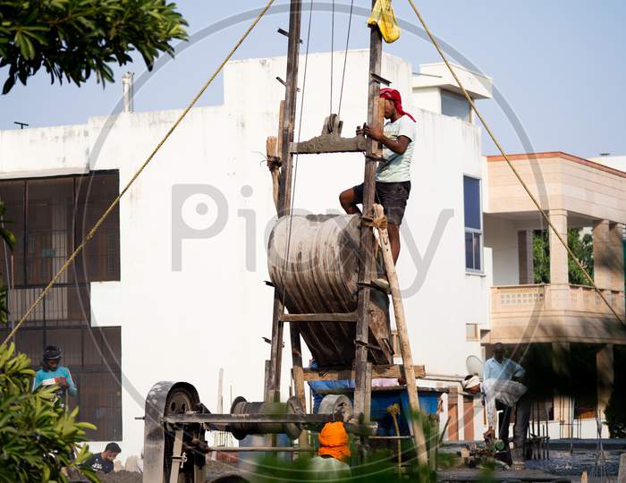 Shot Of Construction Labour With No Safety Equipment Climbing Onto A Hopper And Hammering It To Clean It Off Concrete And Empty It Into A Wagon At An Indian Construction Site