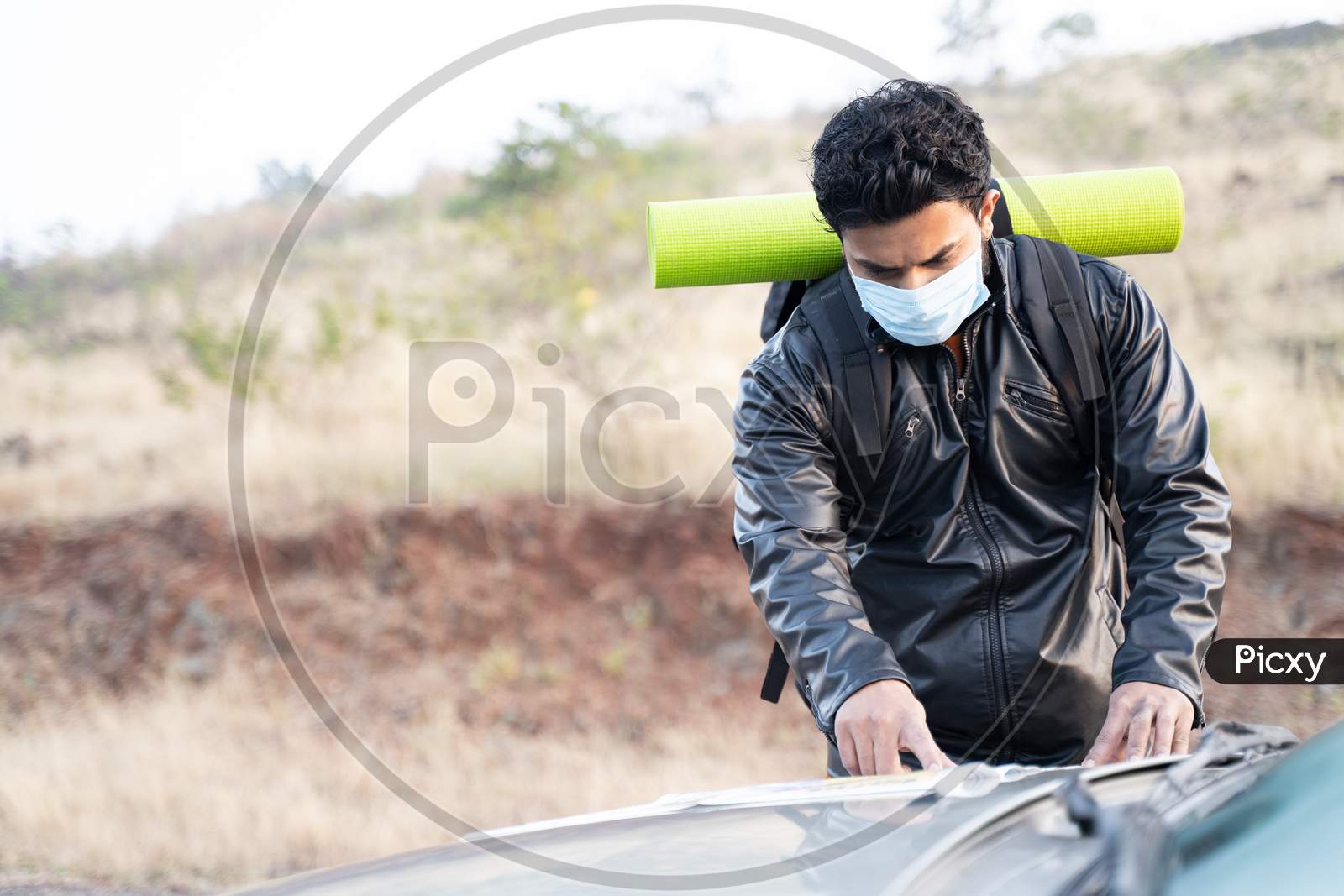 Young Man In Face Mask Busy Searching Or Checking Map On Car Before Starting The Hiking - Concept Of Solo Travel With Safety Measures Due To Coronavirus Covid-19 Pandemic.