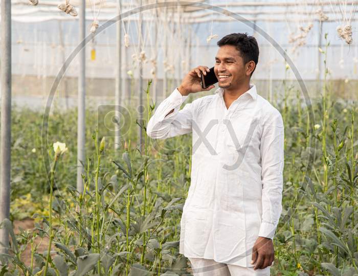 Happy Young Indian Farmer Talking On Smart Mobile Phone At Poly House Or Greenhouse Field, Agriculture And Technology Concept Copy Space