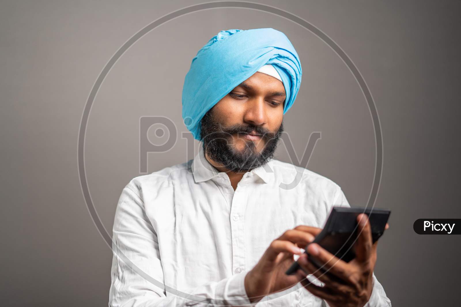 Studio Shot Of Young Sikh Man Seriously Thinking And Calculating Tax Or Financial Expenses On Calculator
