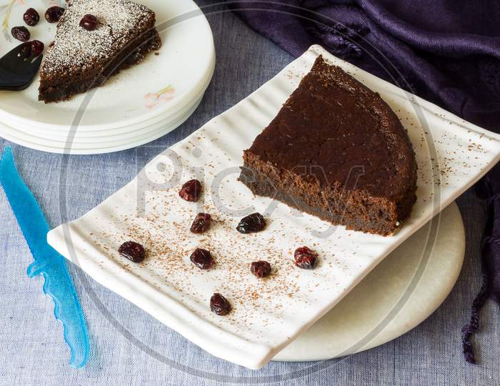 Two slices of dark chocolate cake with cranberry topping and dusting of icing sugar on top, served in white ceramic plates.Top view of delicious cake with knife and purple napkin tucked in the corner