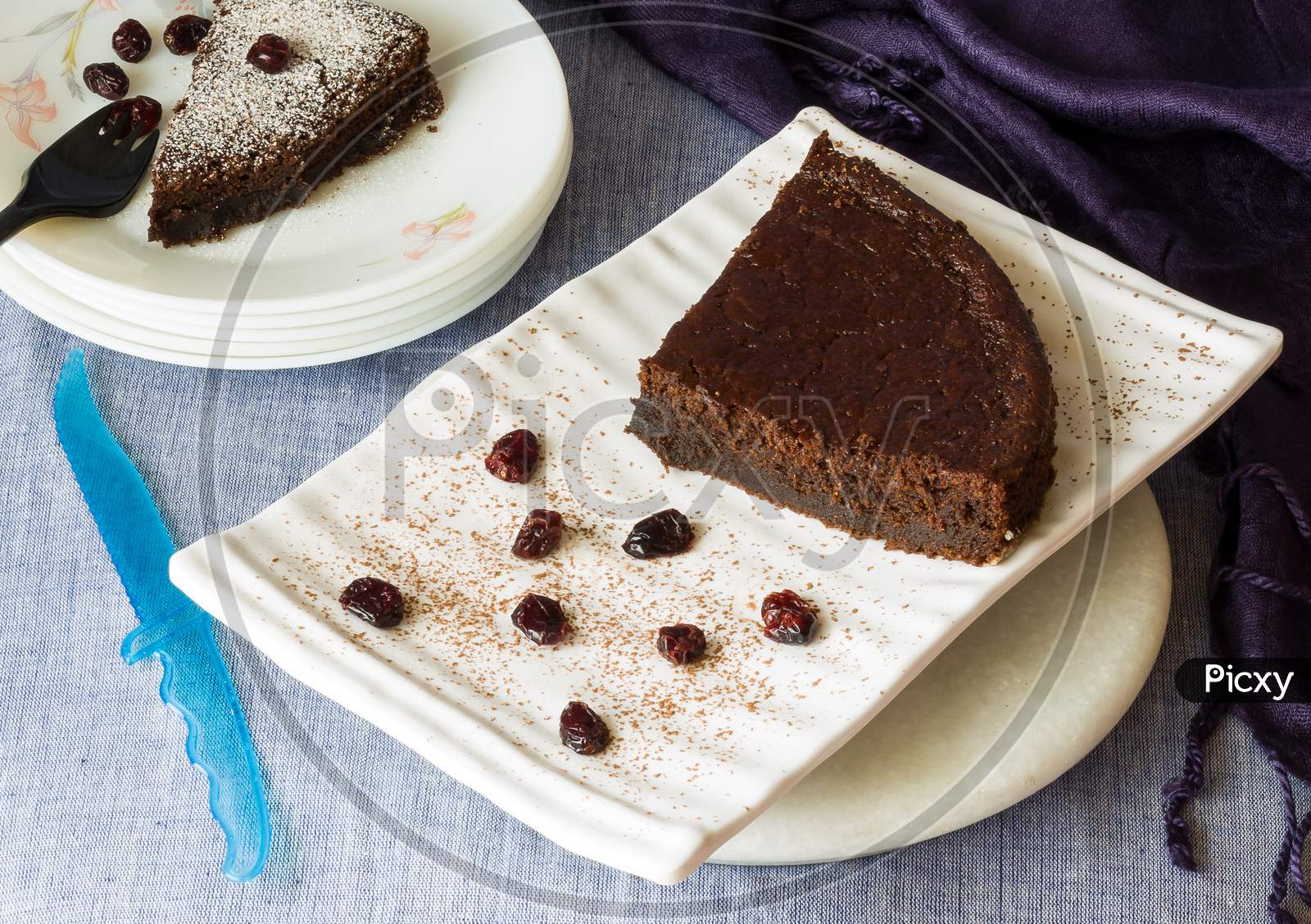 Two slices of dark chocolate cake with cranberry topping and dusting of icing sugar on top, served in white ceramic plates.Top view of delicious cake with knife and purple napkin tucked in the corner