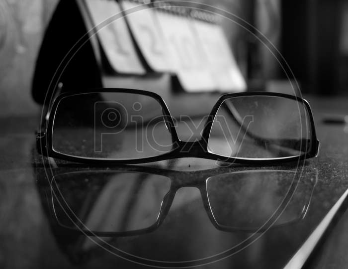 Black And White Image Of Senior Citizen Spectacle Kept On Glass Table Full Of Dust.