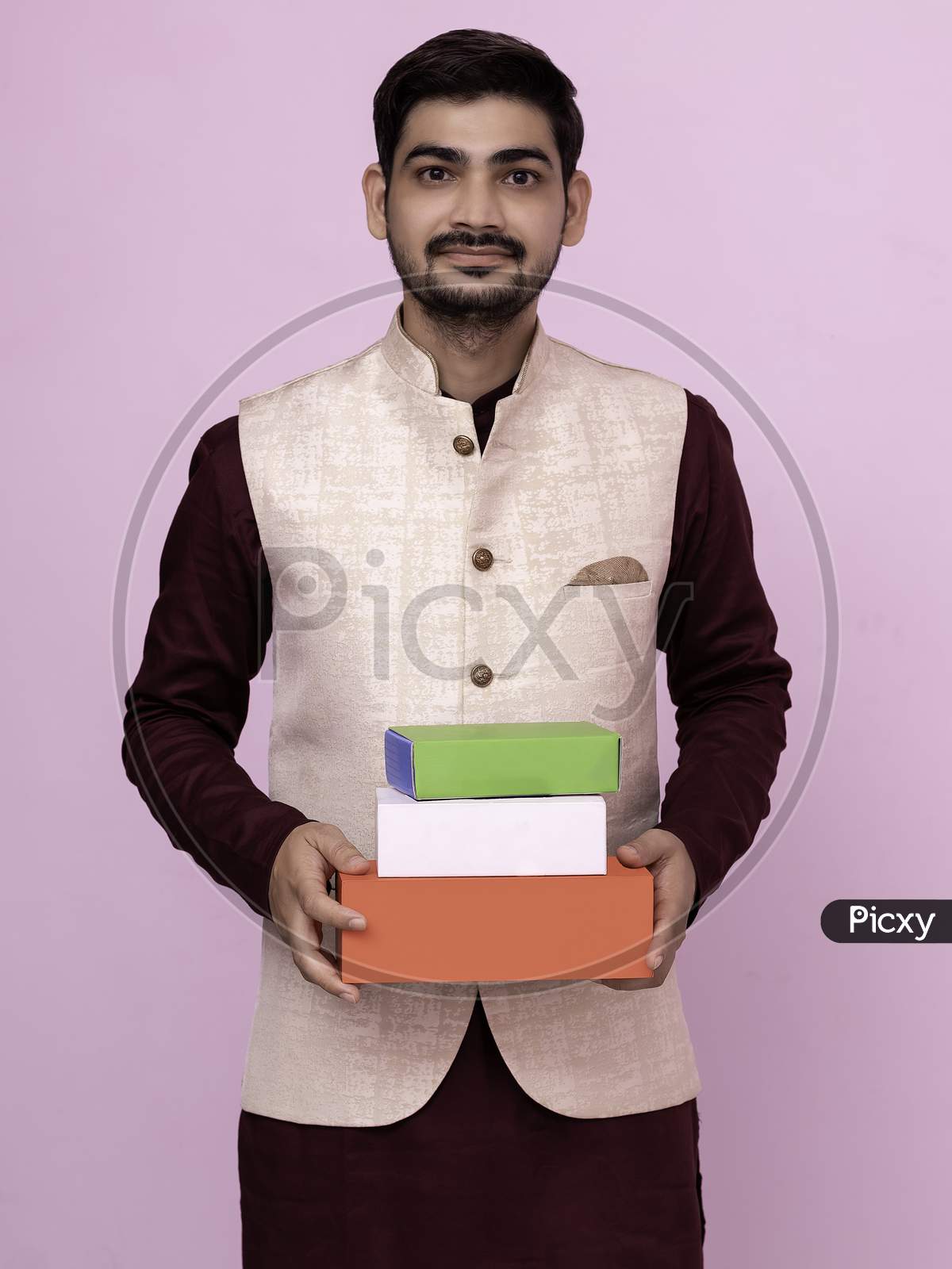 Handsome Indian Man In Traditional Wear Holding Gift Boxes In Hand In Greeting Pose On Isolated Background.