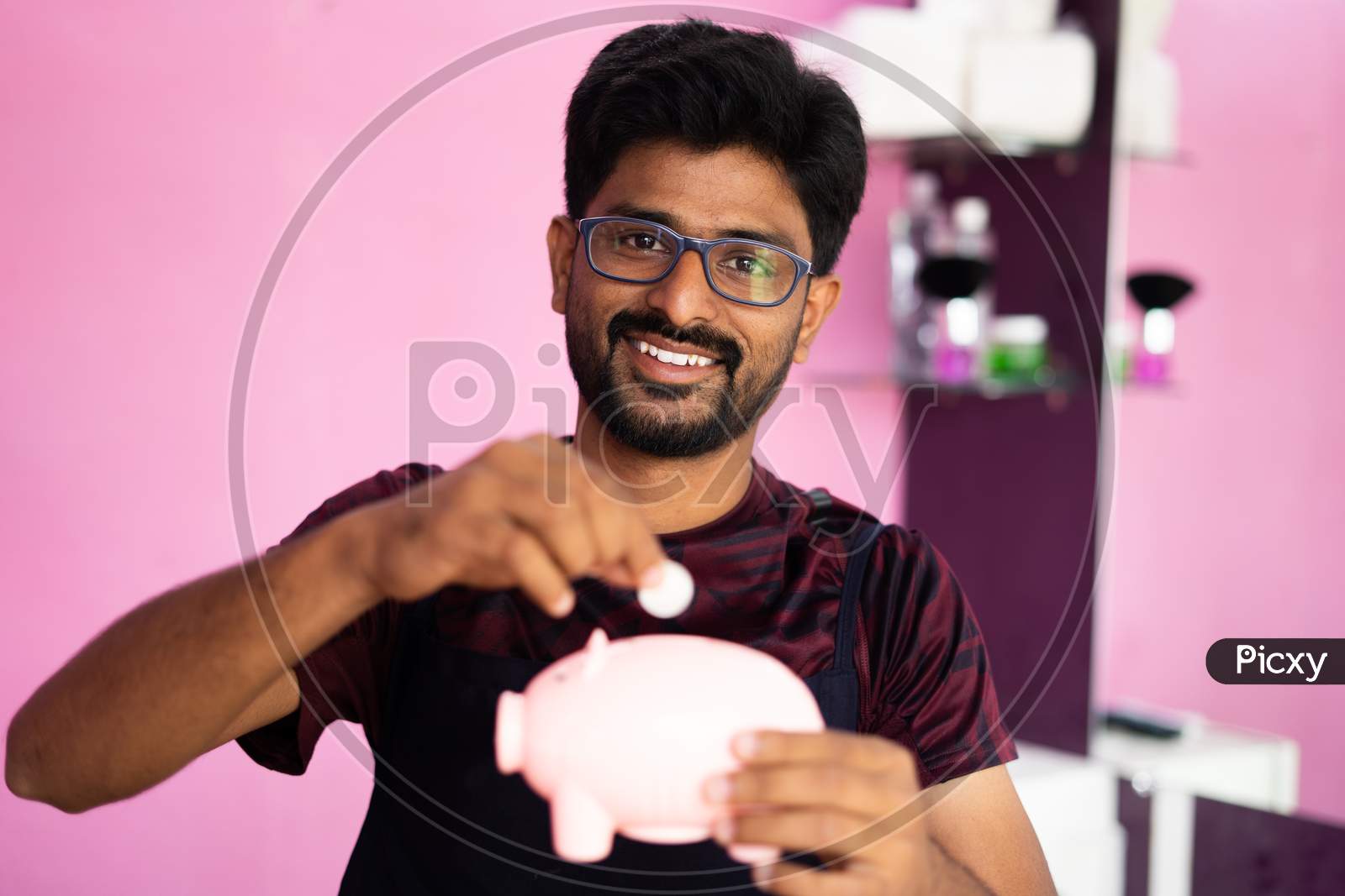 Happy Young Professional Barber Placing Coins Inside Piggy Bank By Looking At Camera - Concept Of Small Business Savings, Financial And Budget.