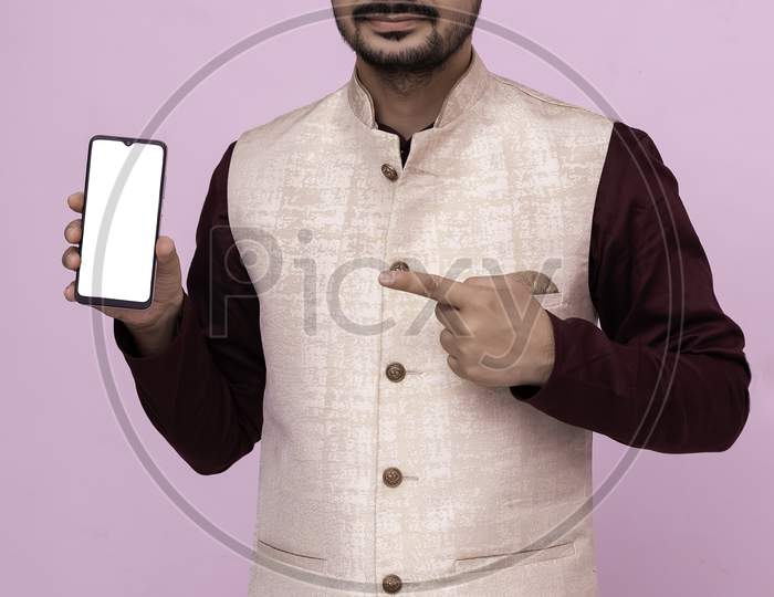 Portrait Of  Man With Beard In Traditional Wear Pointing At Cell Phone And Smiling At Camera. Young Indian Man In Traditional Wear And Showing Smartphone.