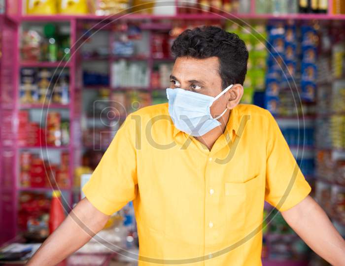 Merchant With Mdical Face Mask At Groceries Or Kirana Store Waiting For Customers By Looking Around During Coronavirus Covid-19 Pandemic - Concept Of Small Businesses Reopen And New Normal Lifestyle.