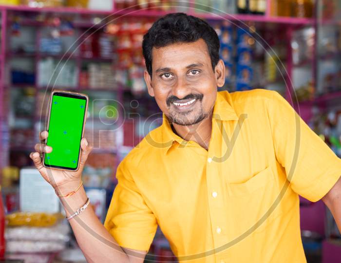 Smiling Merchant At Groceries Store Hold Mobile With Green Screen Mock Up By Looking At Camera - Concept Of Technology, Advertisement, Online Booking And E-Commerce