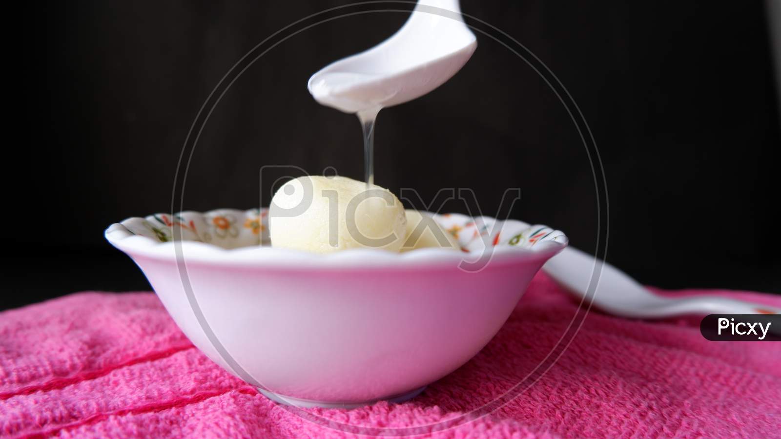 Rasgulla served in white printed bowl and pouring sugar syrup on it by white spoon