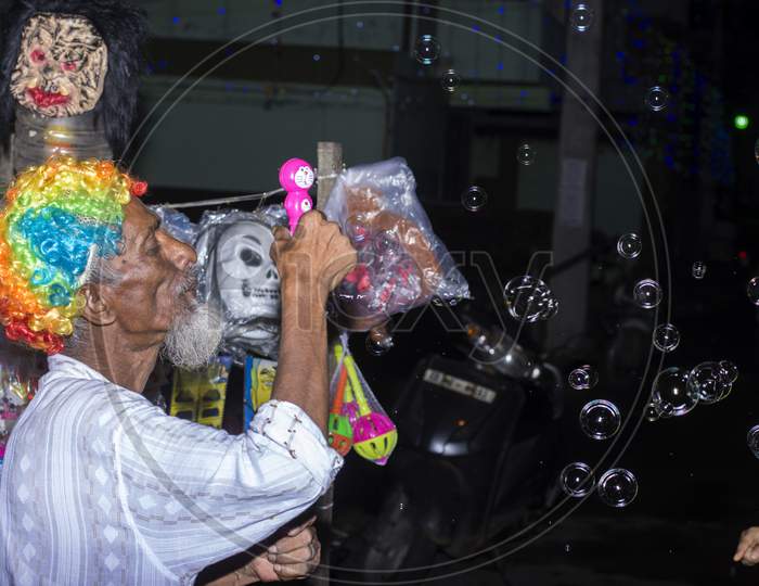 An Old Hawker Wearing Colorful Selling Toys And Creating Bubbles.