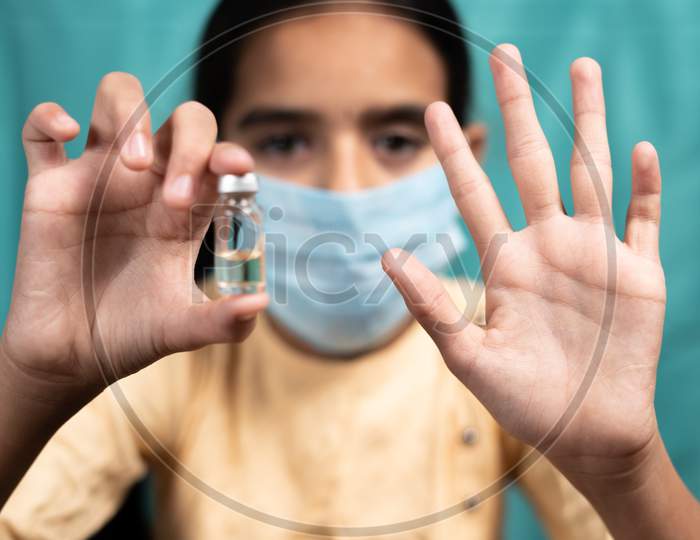 Selective Focus On Hand, Kid Medical Face Mask Saying No To Covid-19 Or Coronavirus Vaccination By Shoing Showing Hand Gesture And Vaccine Bottle - Concept Of Children Vaccine Hesitancy.