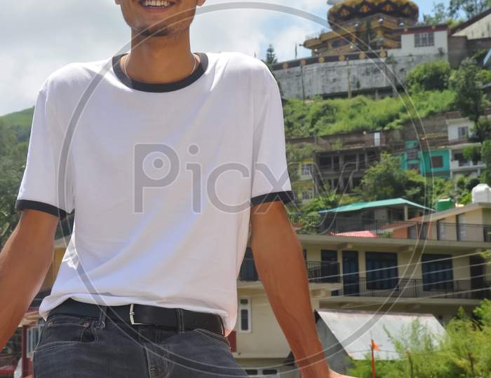 A young guy smiling sitting on safety barrier beside of lake with looking away and beautiful hilly area of Tso-Pema and statue of Padmasambhava (Guru Rinpoche) in background