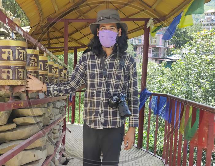 A good looking long haired young guy wearing face mask and sun hat, carrying DSLR camera with turning Buddhist prayer wheel in Tso-Pema with looking at camera
