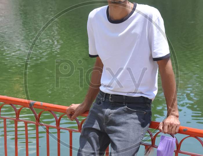A handsome North Indian young guy wearing white t-shirt and black jeans and holding face mask on hand with sitting on safety barrier by lake and looking at camera