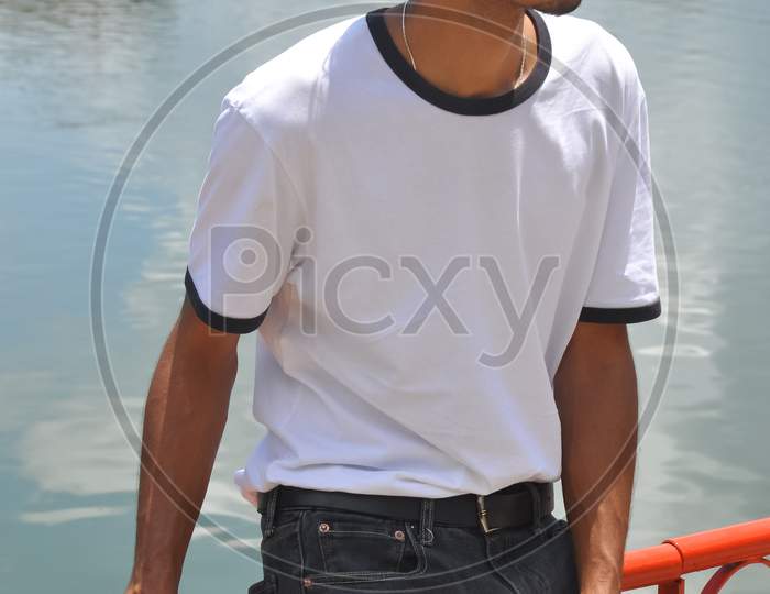 A good looking Indian young guy wearing white t-shirt and black jeans with sitting on safety barrier by lake and looking sideways