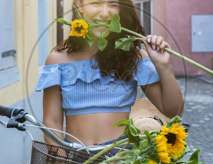 Beautiful Young Lady Biting In Sunflower While Walking Through The Old City And Pushing Her Turquoise Bicycle. Sunbeam At Background.