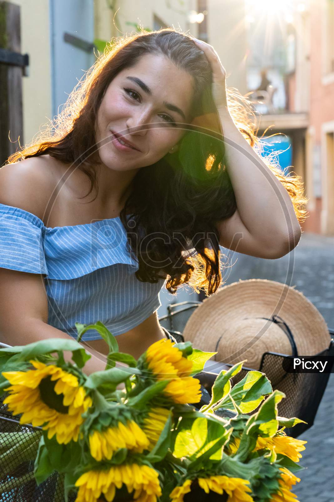 Beautiful Young Lady Walking Through The Old City While Pushing Her Turquoise Bicycle With Sunflowers In A Basket. Sunbeam At Background.