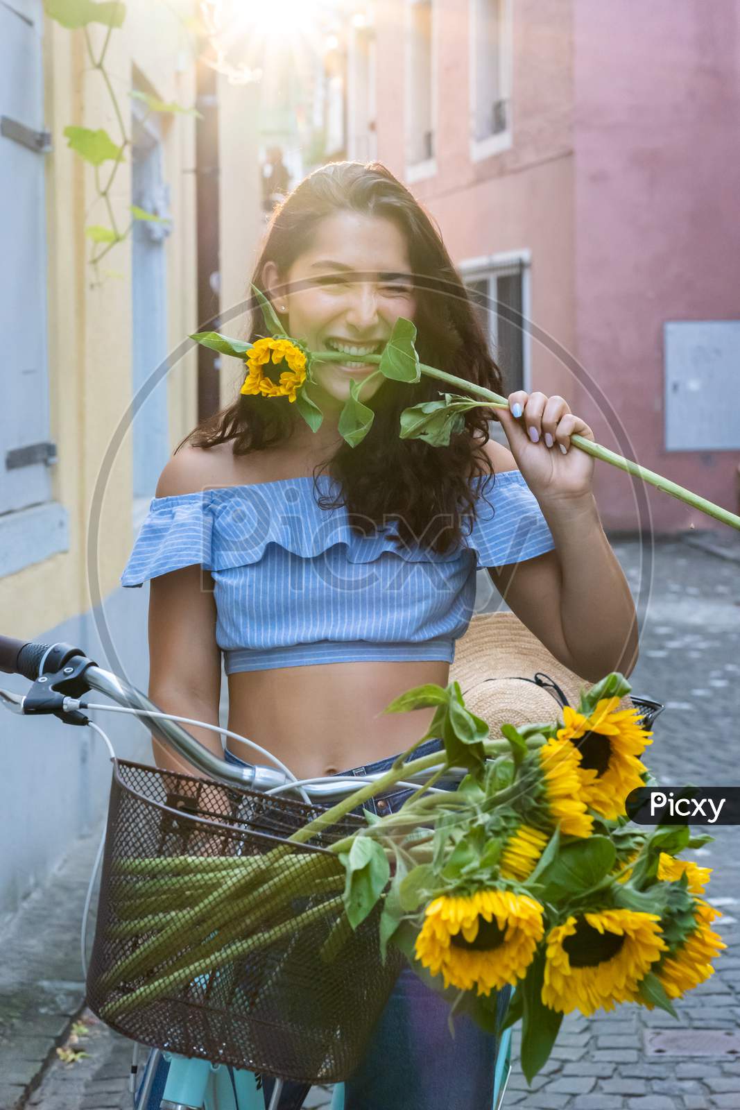 Beautiful Young Lady Biting In Sunflower While Walking Through The Old City And Pushing Her Turquoise Bicycle. Sunbeam At Background.