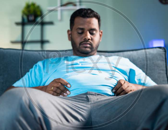 Sad Obese Man Disappointed For Heavy Belly Fat By Holding Hands While Sitting On Sofa At Home - Concept Of Overweight, Disease, Suffering From Excess Stomach Waist And Unhealthy Eating Problem