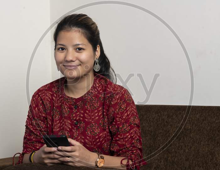 Portrait Of A Young Indian Girl Using Her Mobile Phone While Looking Into The Camera And Smiling.