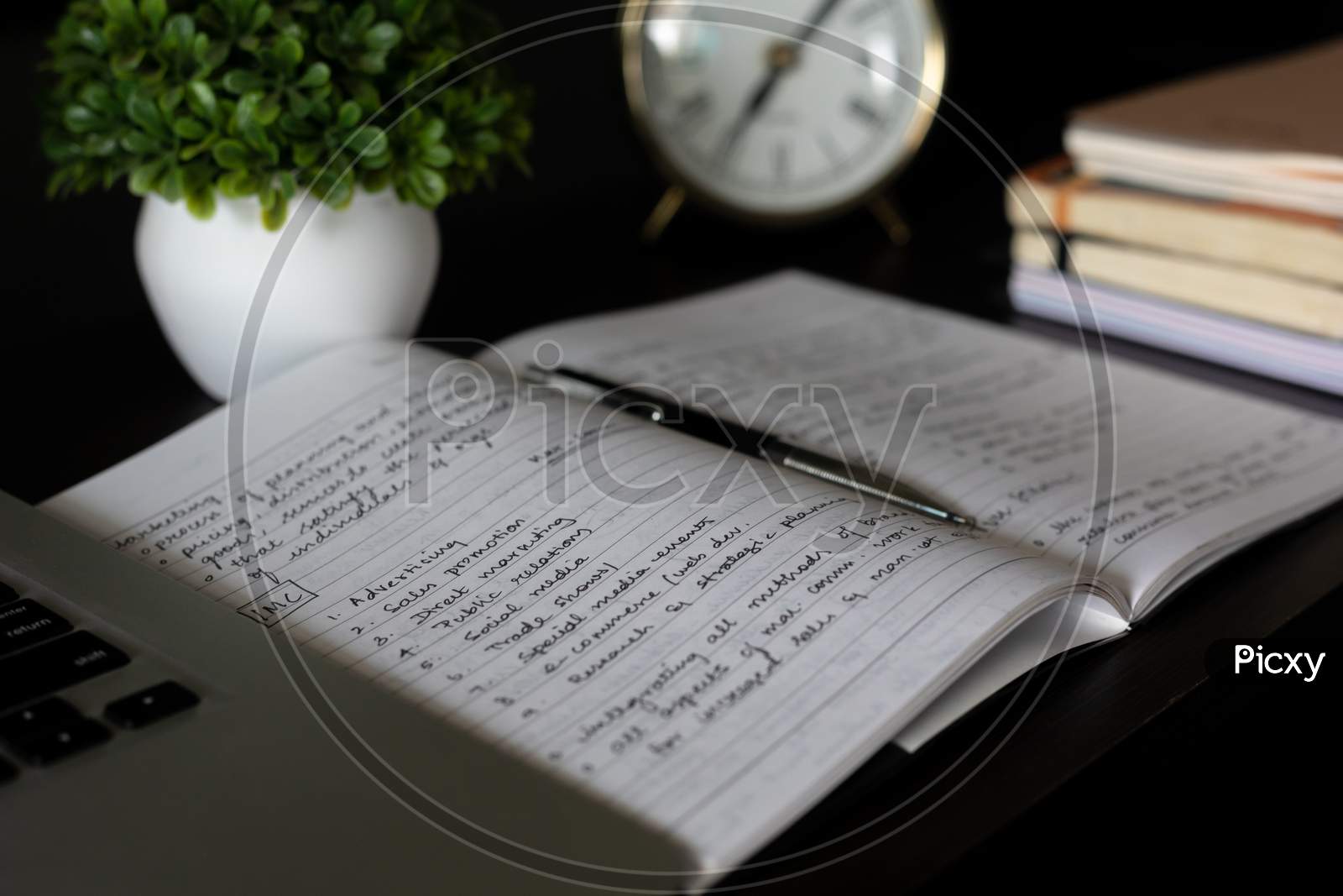 Aesthetic Antique clock showing 7'o clock. Work from home WFH. Workspace with books and indoor plant on dark black office table desk. Closeup, profile shot. Notepad taking notes. Home office bookshelf