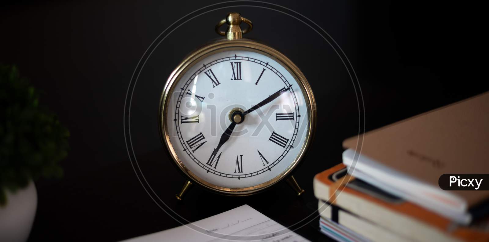 Aesthetic Antique clock showing 7'o clock. Work from home WFH. Workspace with books and indoor plant on dark black office table desk. Closeup, profile shot.