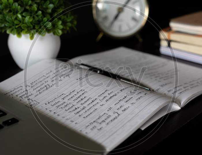 Aesthetic Antique clock showing 7'o clock. Work from home WFH. Workspace with books and indoor plant on dark black office table desk. Closeup, profile shot. Notepad taking notes. Home office bookshelf