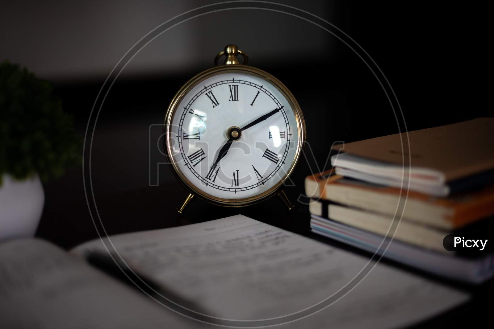 Aesthetic Antique clock showing 7'o clock. Work from home WFH. Workspace with books and indoor plant on dark black office table desk. Closeup, profile shot. Notepad taking notes. Home office.