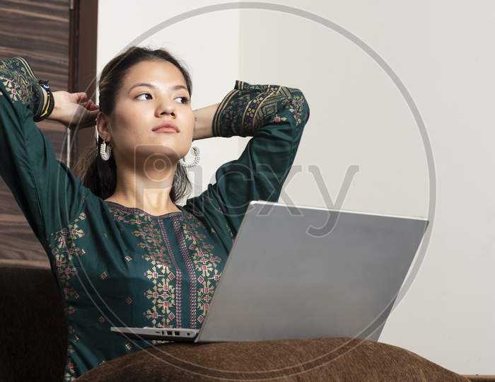Young Indian Girl Working On Her Laptop While Lying Down On The Couch.Smiling Hindu Female Professional Employee Typing Email On Laptop At Workplace