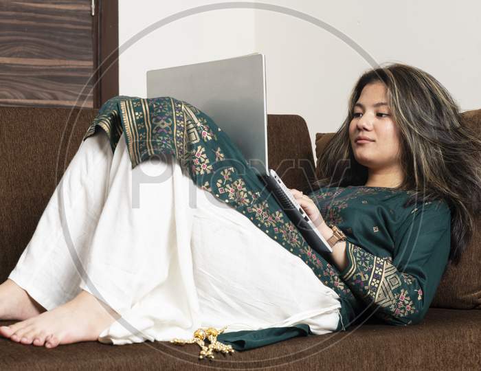 Young Indian Girl Working On Her Laptop While Lying Down On The Couch.Smiling Hindu Female Professional Employee Typing Email On Laptop At Workplace