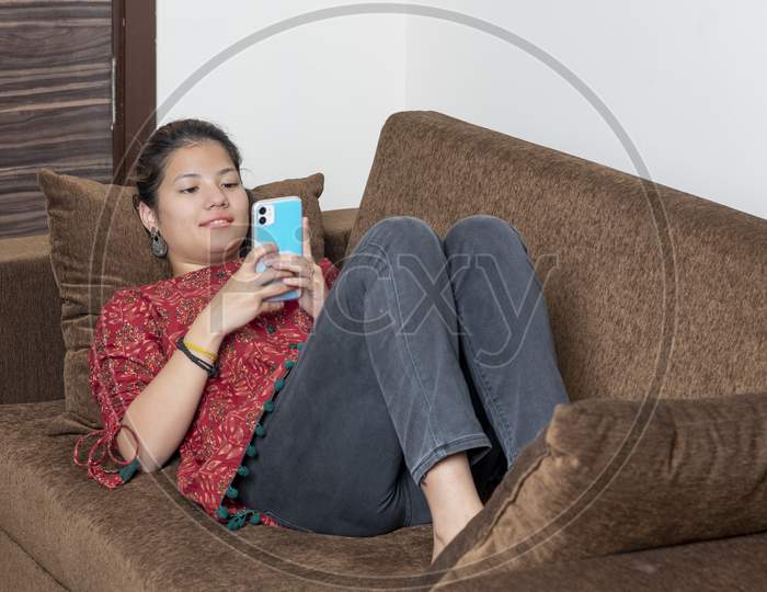 Young Indian Girl Using Her Mobile Phone While Lying On The Couch. Young Generation Using Mobile Phone. Mobile And Technology Concept.