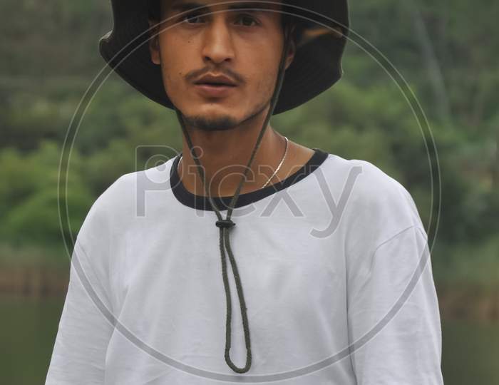 Waist-up shot of a good looking Indian young guy standing outdoor with wearing boonie hat and white tshirt and looking at camera