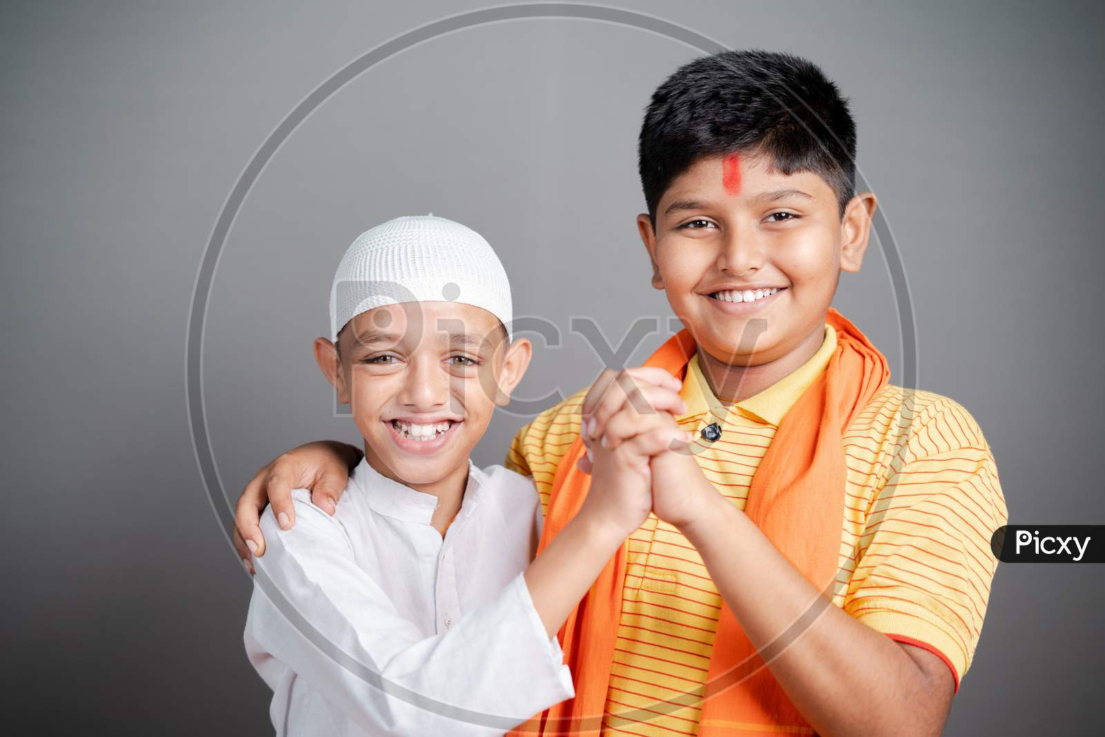 Happy Hindu Muslim Kids Showing Unity By Holding Hands Together By Looking At Camera On Gray Background - Concept Of Diversity, Bonding And Multiethnic Friendship.