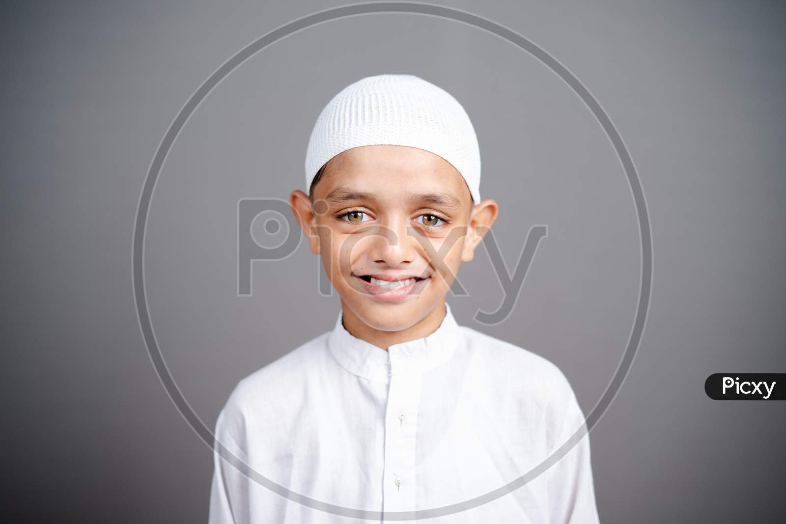 Smiling Muslim Child With Traditional Cap By Looking At Camera On Gray Background - Concept Showing Of Happiness, Positive Emotion During Childhood