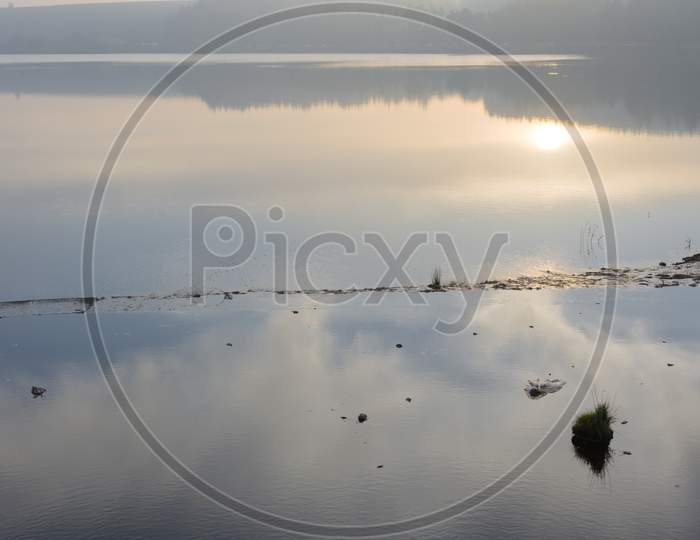 Vertical Hazy Sunset Reflected In Calm Water With Edge Of Reservoir