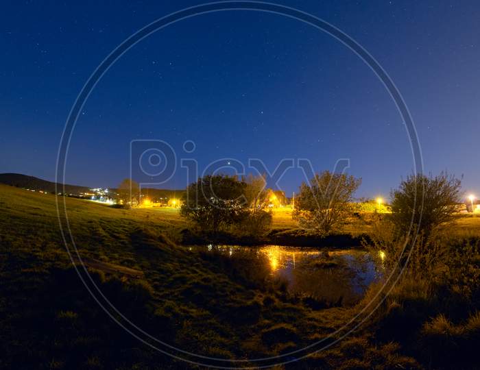 Blue Starry Sky Reflected In Small Pond Contrasts To Line Of Orange Streetlights. Rural Setting Near Hills.