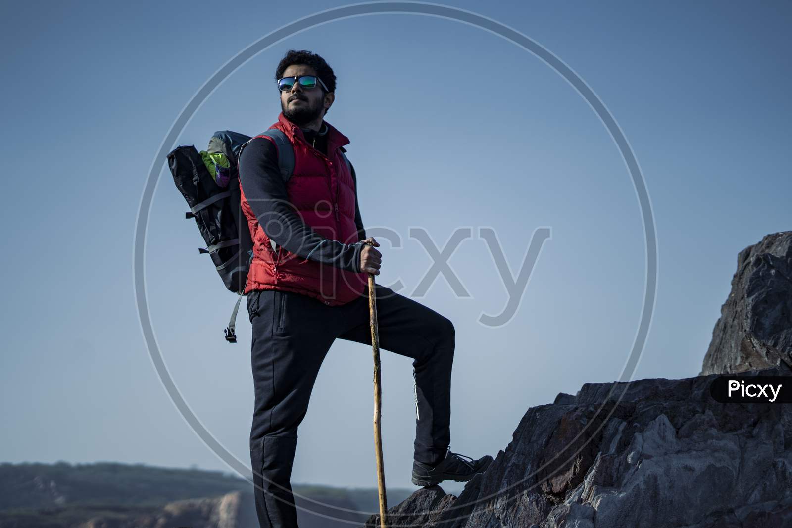 Young Indian Traveler Hiking Up The Mountain With A Backpack And A Stick, With A Beautiful Lake View In The Background. Sports And Freedom Concept.