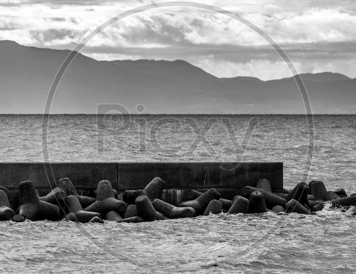 Grayscale Shot Of Tetrapods On A Dock By The Sea Under A Cloudy Sky