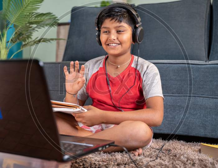 Young Kid With Headphones And Books In Handphone Greeting His Tutor During Online Class On Laptop At Home - Concept Of Virtual Educatiom, Homeschooling And New Normal During Coronavirus Pandemic.