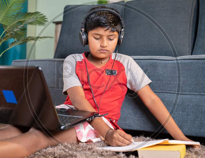 Kid With Headphone Noting Down To Book From Laptop During Virtual Class From Laptop At Home - Concept Of Online Classroom, Online Education, Technology And Lifestyle.