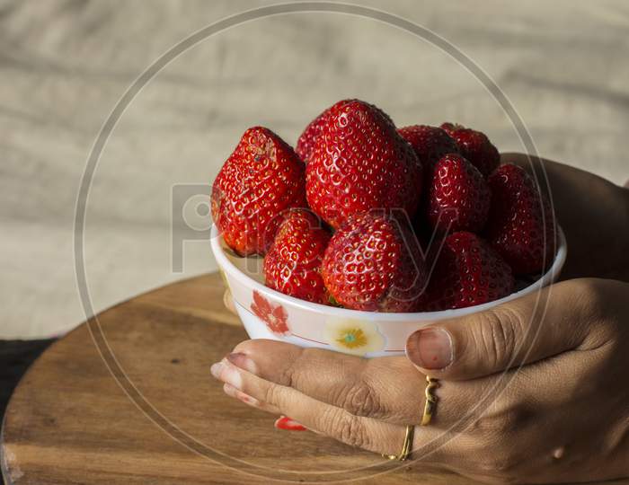 A Female Hand Holding Fresh Organic Red Strawberry In A Bowl On Wooden Table.