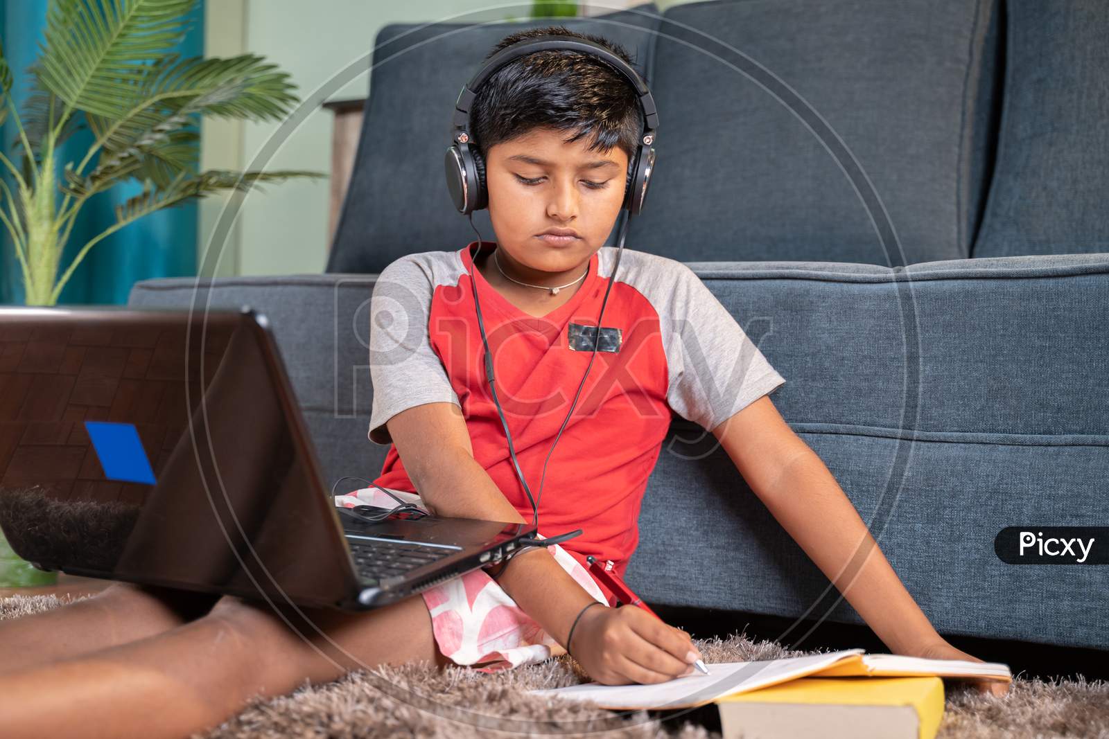 Kid With Headphone Noting Down To Book From Laptop During Virtual Class From Laptop At Home - Concept Of Online Classroom, Online Education, Technology And Lifestyle.