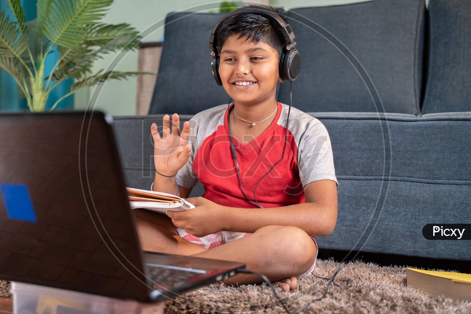 Young Kid With Headphones And Books In Handphone Greeting His Tutor During Online Class On Laptop At Home - Concept Of Virtual Educatiom, Homeschooling And New Normal During Coronavirus Pandemic.