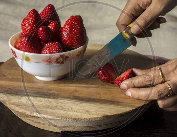 A Female Hand Cutting Fresh Organic Red Strawberry In A Bowl On Wooden Table.