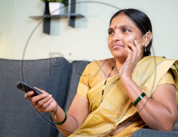 Serious Indian Woman In Saree Watching Tv Serials At Home While Sitting On Sofa By Holding Remote During Leisure Time