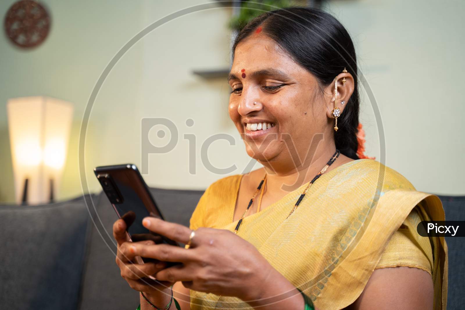 Happy Smiling Indian Woman Busy Using Mobile Phone While Sitting On Sofa - Concept Of People Using Social Media, Online Messaging App, Internet And Entertainment Lifestyle.
