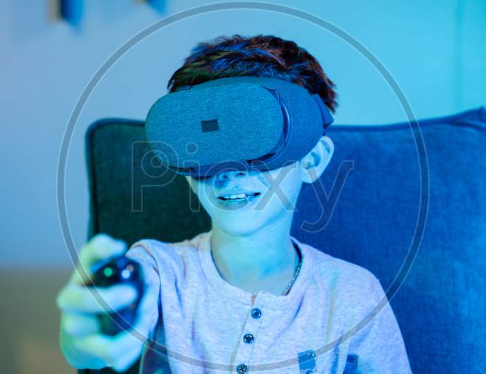 Young Kid Playing Game By Wearing Vr Or Virtual Reality Headset And Game Pad While Sitting On Sofa Showing With Neon Light Effect.