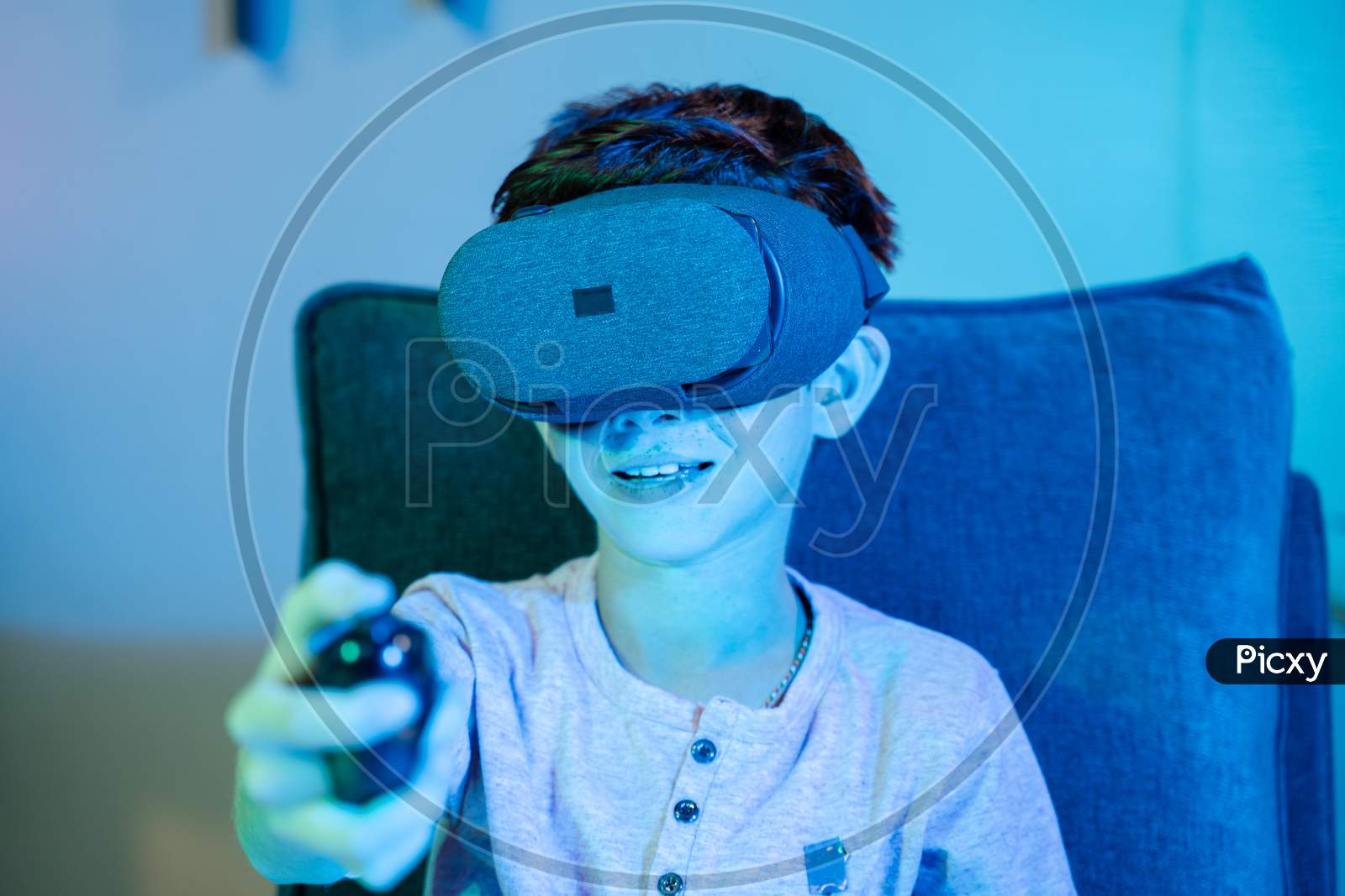 Young Kid Playing Game By Wearing Vr Or Virtual Reality Headset And Game Pad While Sitting On Sofa Showing With Neon Light Effect.