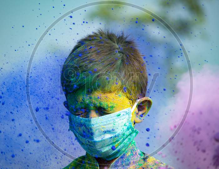 Portrait Shot Of Young Kid With Face Mask Standing While Colorful Holi Powder Throwing At Him - Concept Of Holi Festival During Coronavirus Coivd-19 Pandemic